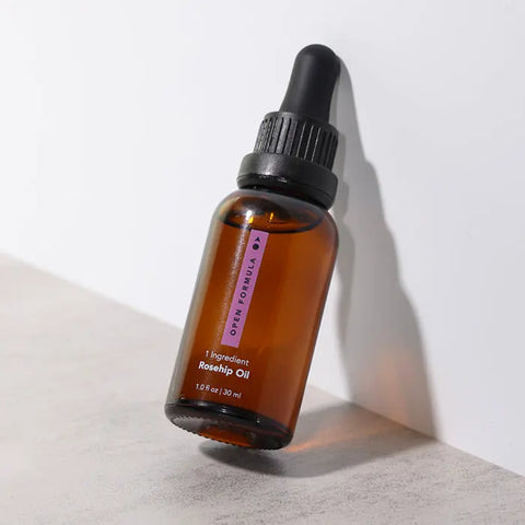 A brown glass dropper bottle of Open Formula Rosehip Oil, labeled 1 Ingredient, with a black cap, sits on a light gray surface against a white wall.