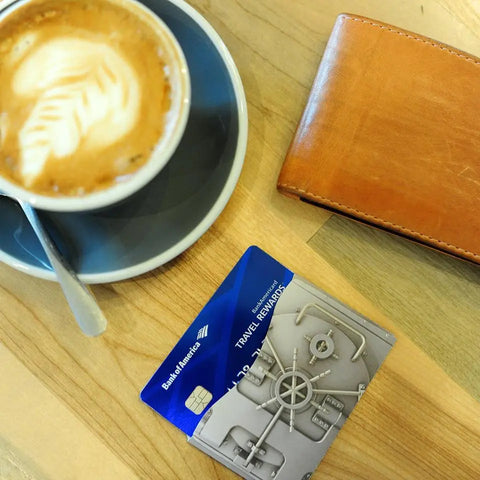 A cup of coffee with a spoon, a brown wallet, and two credit cards—one Bank of America Travel Rewards—plus Rogue Industries RFID Blocking Credit Card Sleeves (Platinum Vault) on a wooden table for enhanced identity theft protection.
