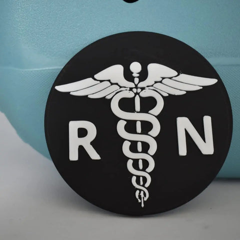 The Boglets RN Nurse Charm by Chatelet Manufacturing features a black circular badge with a white caduceus and RN letters, ideal nurse gifts or stylish decorative charms compatible with Bogg Bags, Simply Southern, and other tote bags.