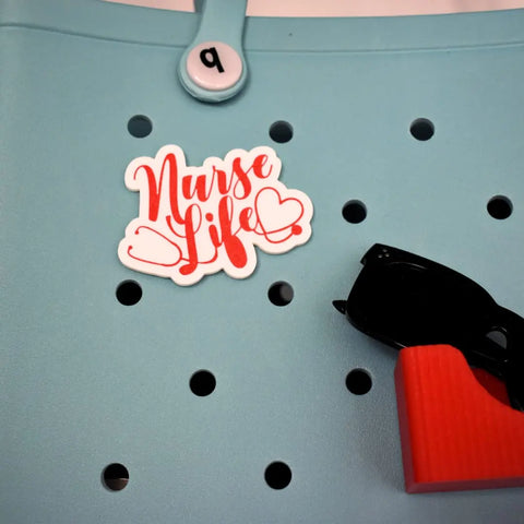 The Chatelet Manufacturing Boglets RN Nurse Charm adds style to any tote with a “Nurse Life” pin, number 9 button, and sunglasses charm—perfect decorative charms for nurses and compatible with Bogg Bags and more.