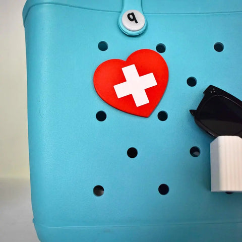 The Boglets RN Nurse Charm by Chatelet Manufacturing, featuring a red heart with a white cross and sunglasses design, is perfect for nurses and easily clips onto Bogg Bags, Simply Southern, or other tote bags for a decorative touch.