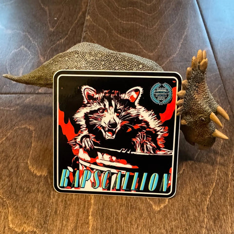 A toy dinosaur displays the American Lictor Sticker: Raccoon - Rapscallion, which features a raccoon with flames in the background and RAPSCALLION in bold at the bottom.
