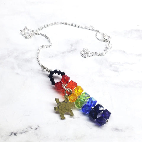 The Wire Woven Rainbow Autism Puzzle Piece Bar Necklace-2024 Limited Edition by Alexa Martha Designs showcases vibrant crystal beads and a puzzle piece charm, beautifully displayed on a white marble surface.