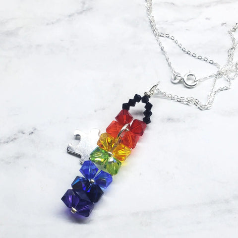 The Wire Woven Rainbow Autism Puzzle Piece Bar Necklace—2024 Limited Edition by Alexa Martha Designs features a vertical bar with rainbow crystals and a silver puzzle piece charm, beautifully displayed on a white marble background.