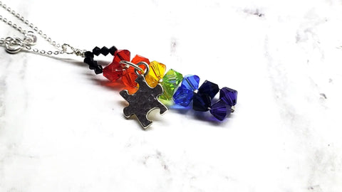 The Alexa Martha Designs Wire Woven Rainbow Autism Puzzle Piece Bar Necklace-2024 Limited Edition features vibrant rainbow beads, a silver puzzle piece charm, and a sleek chain, elegantly showcased on white marble.