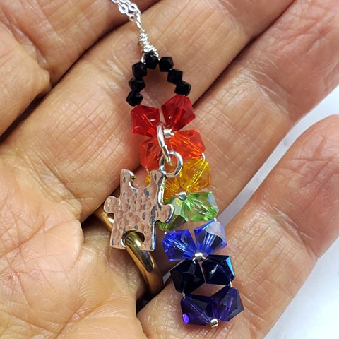 Close-up of a hand holding the Wire Woven Rainbow Autism Puzzle Piece Bar Necklace-2024 Limited Edition by Alexa Martha Designs, featuring a silver puzzle piece and faceted rainbow beads accented with black beads.