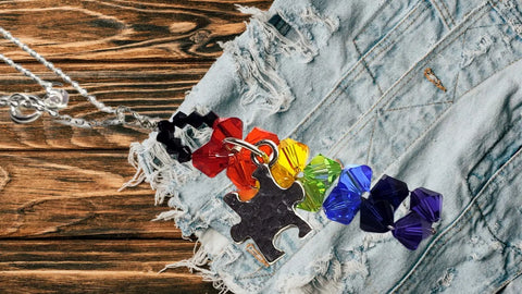 The Wire Woven Rainbow Autism Puzzle Piece Bar Necklace-2024 Limited Edition by Alexa Martha Designs features a rainbow puzzle piece bar, displayed on frayed light blue denim over wood, symbolizing autism awareness.
