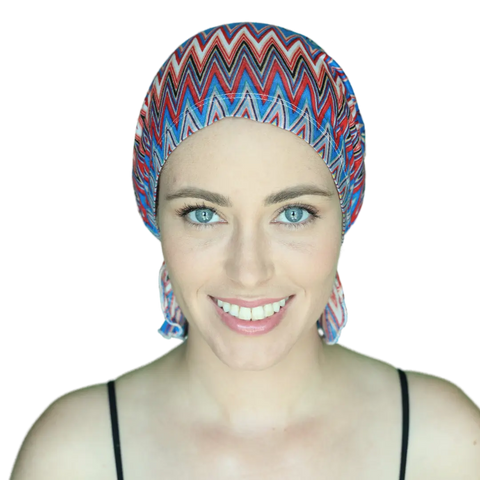 A smiling woman with bright blue eyes wears the Chemo Beanies® Louisa zigzag-patterned chemo beanie and a black spaghetti strap top, looking directly at the camera against a plain background.