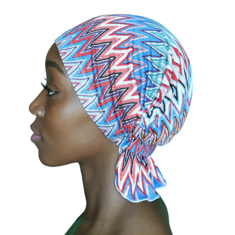 A woman in profile wears the Chemo Beanies® Louisa chemo scarf, featuring a colorful zigzag pattern in blue, red, and white, tied at the back of her head. The background is plain black.