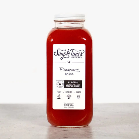 A clear bottle of Simple Times Mixers Raspberry Mule sits on a gray surface, its label noting the mule cocktail mixer is handcrafted with cold-pressed raspberries and all-natural ingredients.