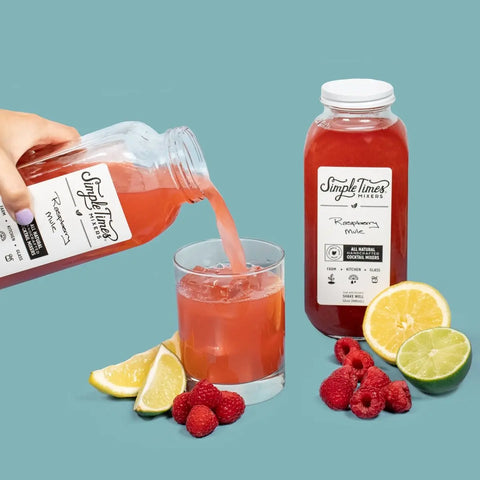 A hand pours Simple Times Mixers Raspberry Mule, made with cold-pressed raspberries, into a glass. Raspberries, lemon and lime slices are on the table beside another Raspberry Mule bottle, set against a light blue background.