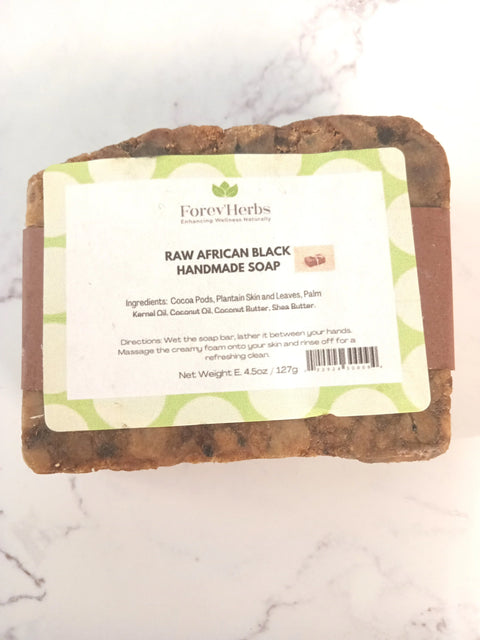 A rectangular bar of Raw African Black Handmade Soap from Forev'Herbs, with a white and green label showing ingredients and directions, rests on a light marble surface. The Forev'Herbs brand name appears clearly on the label.