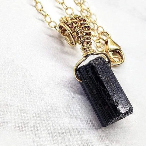 The Raw Black Tourmaline Gemstone Necklace by Alexa Martha Designs features a raw black tourmaline crystal pendant on a gold filled chain with a lobster clasp, displayed on a white surface.