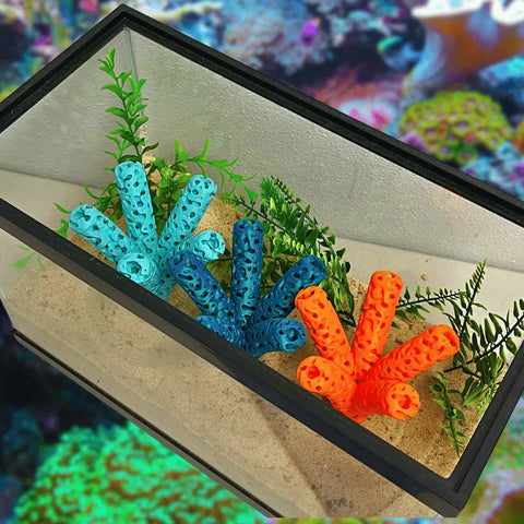 The Chatelet Manufacturing Aquarium Coral Frag Stand Branch, featuring a lattice coral structure and 5 frag mounts, is displayed in a rectangular aquarium with sand, green artificial plants, and blue-orange coral decor against an underwater-themed background.