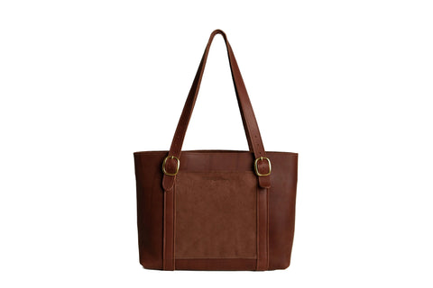The Regina Tote by Urban Southern is a handcrafted brown leather bag with dual shoulder straps, gold-tone buckles, and a spacious front pocket, showcased against a white background.