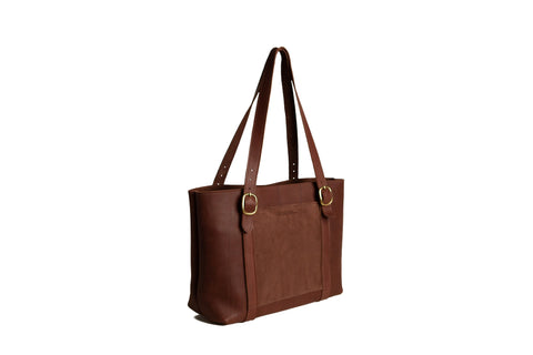 The Regina Tote by Urban Southern is a brown full-grain leather bag with two long shoulder straps and gold buckle accents, displayed at an angle against a white background.