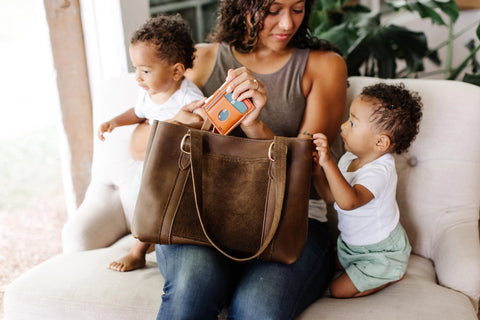A woman sits on a chair with two young children—one on her lap, one standing—both in white shirts. She holds Urban Southerns Regina Tote full-grain leather bag and an orange wallet, looking down at the kids.
