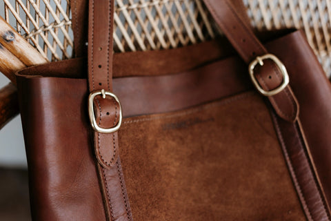 Close-up of the Urban Southern Regina Tote, a handcrafted brown suede leather bag with gold buckles, hanging on a woven chair.