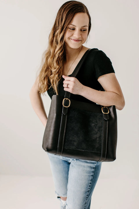 A woman with long, wavy brown hair in a black shirt and ripped light blue jeans smiles while holding the Regina Tote by Urban Southern over her shoulder against a plain white background.