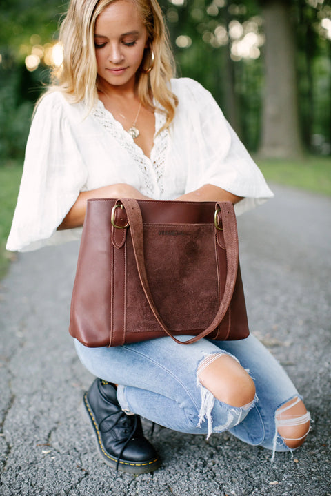 A woman with blonde hair kneels on a gravel path, looking into the Urban Southern Regina Tote. She wears a white blouse and ripped jeans, with trees and greenery forming a natural backdrop.