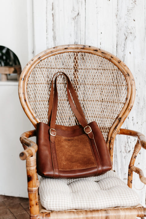The Urban Southern Regina Tote, a handcrafted leather bag with suede accents, rests on a cushioned rattan chair before a rustic white wooden wall.
