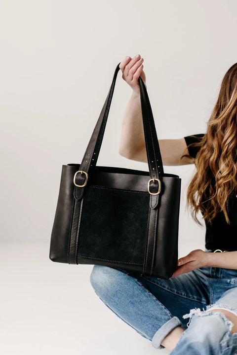 A person with wavy hair, in a black shirt and ripped jeans, sits on the floor holding up the Urban Southern Regina Tote—a handcrafted leather bag with gold buckles—against a plain white background.