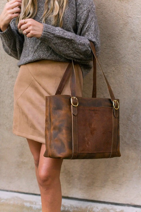 A person with long blonde hair wears a gray sweater and tan suede skirt, holding the Regina Tote by Urban Southern—handcrafted leather with gold buckle accents—while standing against a textured beige wall.