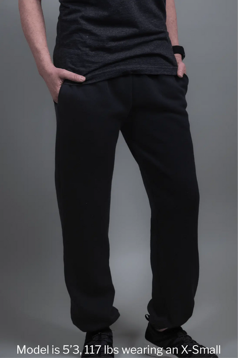 A person stands against a grey background wearing Proudly USA Relaxed Fit Fleece Sweatpants (Black) and a dark t-shirt, hands in pockets. Model is 5’3, 117 lbs, wearing X-Small—perfect for comfy style lovers.