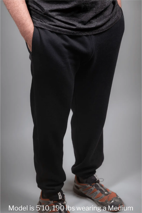 A person wears Proudly USA Relaxed Fit Fleece Sweatpants (Black) with brown sneakers, hands in pockets. Text: Model is 510, 190 lbs, wearing Medium. Comfy clothes made in USA.