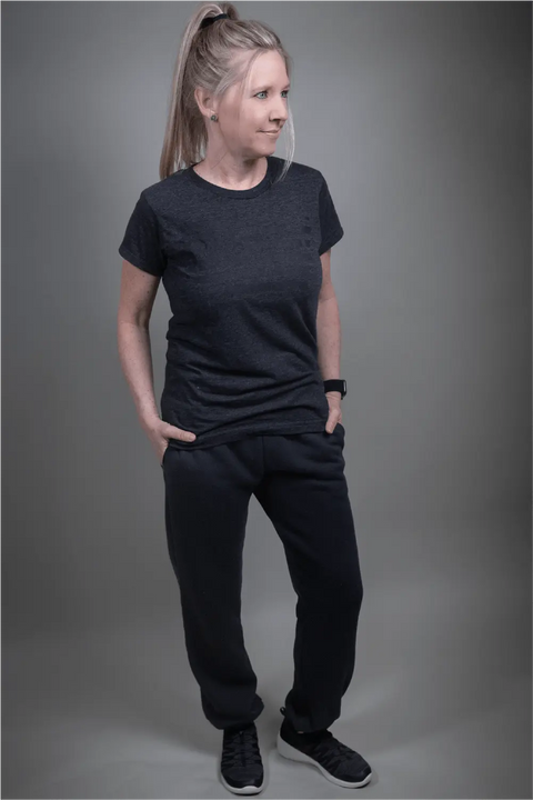 A woman with long blonde hair in a ponytail stands with her hands in her pockets, wearing Proudly USAs Relaxed Fit Fleece Sweatpants (Black) and a black t-shirt, posing against a plain gray background.