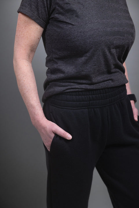 A person stands with one hand in their pocket against a plain gray background, wearing black Proudly USA Relaxed Fit Fleece Sweatpants and a short-sleeve dark gray t-shirt. Their face is not visible.