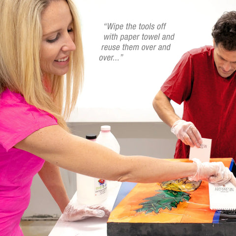 A smiling woman and a man in gloves use the ArtResin Accessory Kit by ArtResin to apply resin to a colorful leaf artwork. Text reads: “Wipe the tools off with paper towel and reuse them over and over…”.