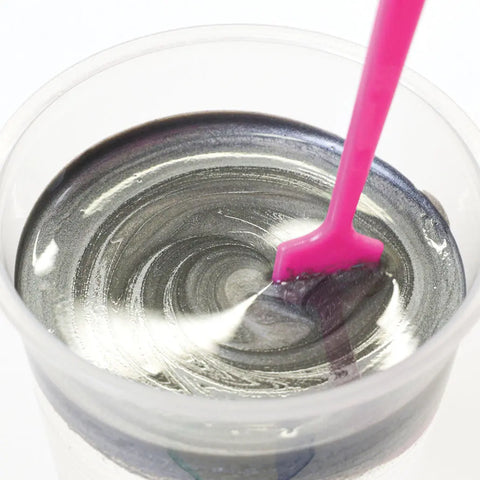 A close-up of ArtResin’s ResinTint Complete Set BUNDLE - 16 Colors in use: swirling silver pigment is mixed in a clear cup with a bright pink spatula, creating a metallic sheen as the liquid reflects light.