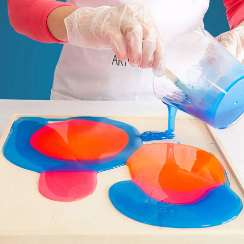 Wearing gloves and a white apron, an artist pours blue from the ArtResin ResinTint Complete Set BUNDLE - 16 Colors onto a wooden board, mixing it with orange and pink tints to create an abstract masterpiece.