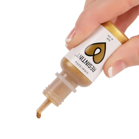 A hand holds an ArtResin ResinTint Metallics bottle upside down, squeezing a drop of gold pigment from the nozzle.