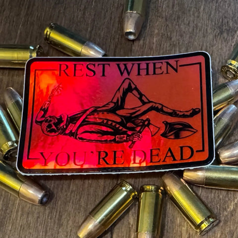 The Sticker: Rest When Dead by American Lictor is a 3 x 2 motivational sticker with a red skeleton and bullets design and the bold text REST WHEN YOURE DEAD, shown on a wooden surface.