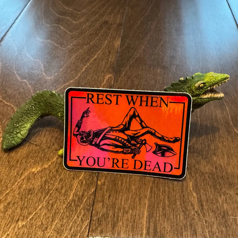 A green plastic dinosaur toy holds an American Lictor Sticker: Rest When Dead (3 x 2) in its mouth, featuring a skeleton lying down with the phrase REST WHEN YOURE DEAD.