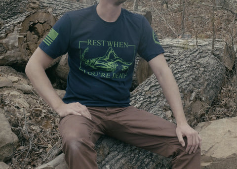 An active individual sits on a fallen tree in the woods, wearing the American Lictor T-Shirt: Rest When Youre Dead, featuring green graphics and a bold motivational message. Their head is out of frame.