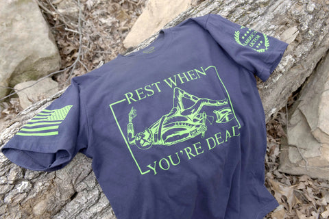 The American Lictor T-Shirt: Rest When Youre Dead features neon green graphics, a skeleton resting, and the slogan REST WHEN YOURE DEAD on navy blue fabric with green sleeve designs—perfect for those who choose to live boldly.