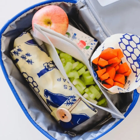 The Lunch Pack by Bee’s Wrap includes an apple, chopped celery, sliced carrots in reusable wraps, plus another item in patterned beeswax cloth, all neatly arranged inside a blue lunch box.