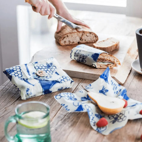With Bees Wrap The Lunch Pack, bread and fruit stay fresh under reusable, washable wraps as someone slices bread on a wooden board. A glass of water and a potted plant create a cozy, eco-friendly vibe.