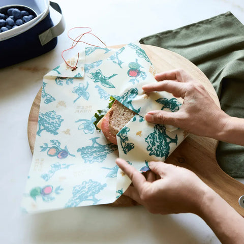 Someone wraps a sandwich using The Lunch Pack by Bees Wrap on a wooden board, beside a green napkin and a container of blueberries.