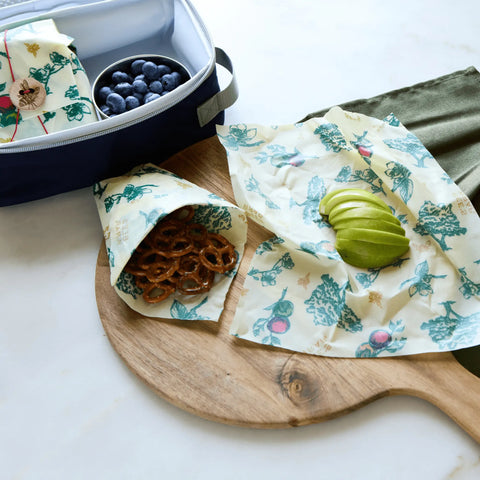 The Lunch Pack by Bees Wrap, featuring reusable wraps with a blue and green veggie print, holds pretzels and sliced green apple—plus a lunchbox with blueberries—all displayed on a wooden board with a green cloth.