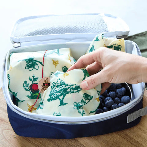 A hand reaches into a lunch box filled with food wrapped in Bees Wrap The Lunch Pack, reusable beeswax wraps featuring green trees and plants. Fresh blueberries peek out, emphasizing these eco-friendly, washable wraps.