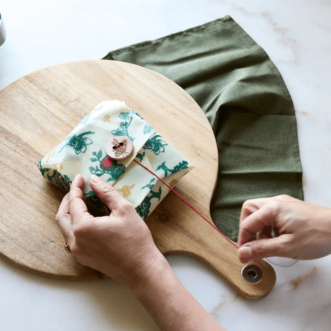 A person wraps a gift with Bees Wrap The Lunch Pack, ties it with red string on a round wooden board, and places an olive green cloth nearby on a white surface.
