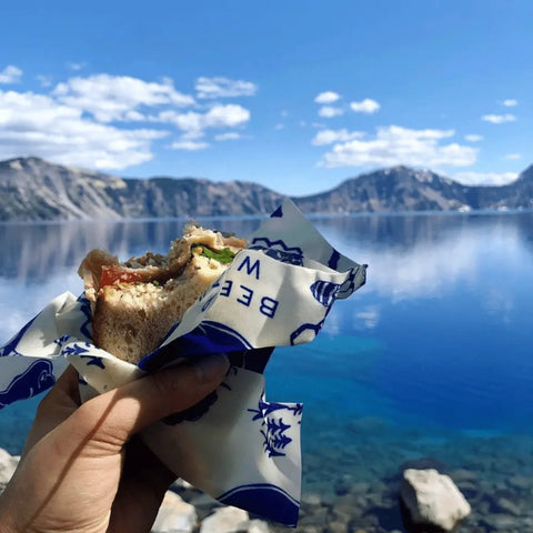 A hand holds a half-eaten sandwich wrapped in Bees Wrap The Lunch Pack, with a clear blue lake and distant mountains under a partly cloudy sky in the background.