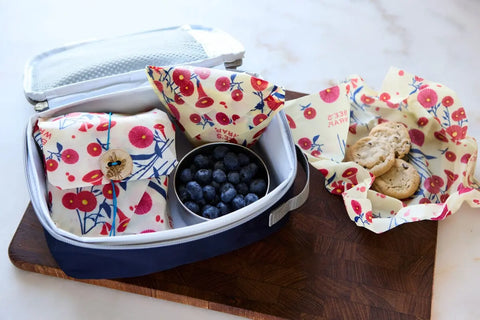 The Lunch Pack by Bees Wrap includes colorful floral reusable wraps, a metal container of blueberries, and cookies on a wooden cutting board. Some cookies are wrapped in matching washable Bee’s Wraps outside the lunchbox.