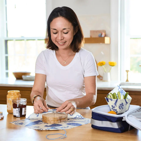 A woman in a bright kitchen smiles while preparing a sandwich. Beside her are the Bees Wrap Lunch Pack, jars of peanut butter and jelly, a lunchbox, washable wraps, and sliced vegetables.