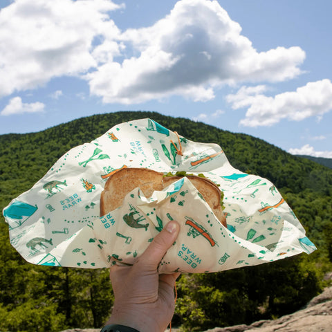 A hand holds a sandwich wrapped in Bee’s Wrap The Lunch Pack compostable wrap, with mountains and trees in the background beneath a partly cloudy sky.