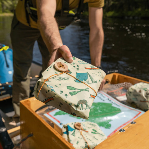 Wearing outdoor gear, someone hands The Lunch Pack by Bee’s Wrap—a small, gift-wrapped package in reusable, nature-themed wraps—from a wooden box on a canoe near a river, with other Bees Wrap packs and a map nearby.